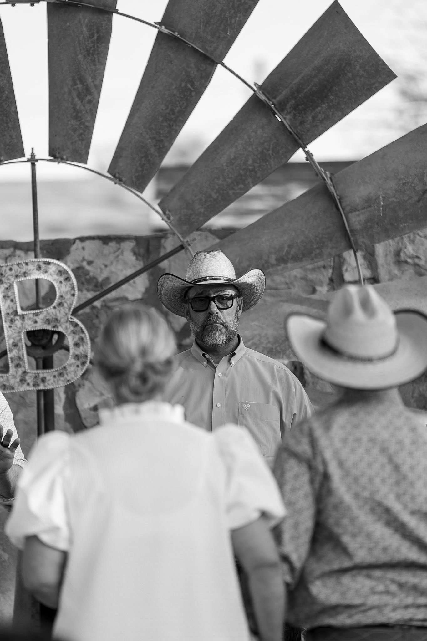 Black and white photo of a bearded man in cowboy hat and sunglasses standing near a windmill at outdoor event.