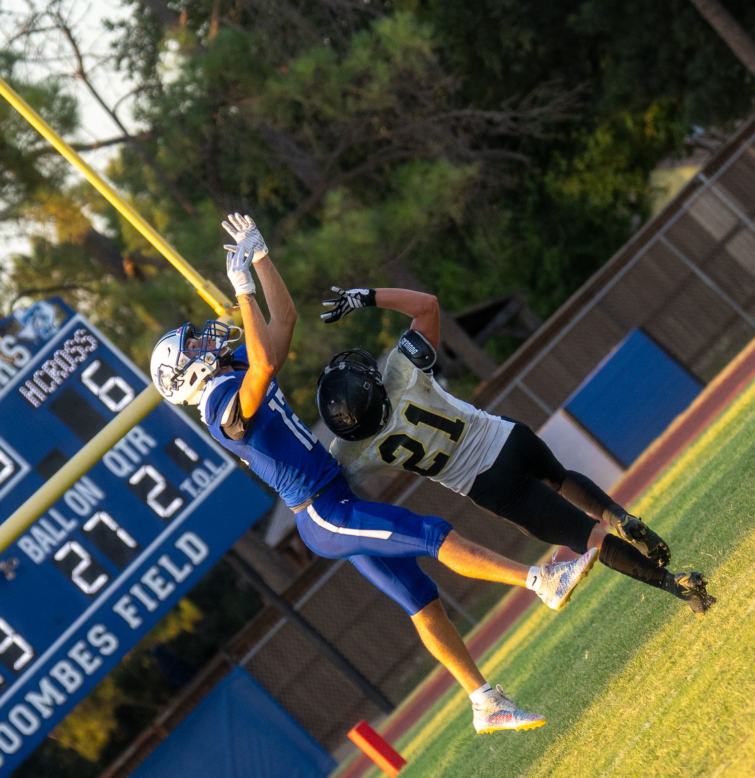 Two football players leap for the ball during a high school game on Dombes Field under bright sunlight.