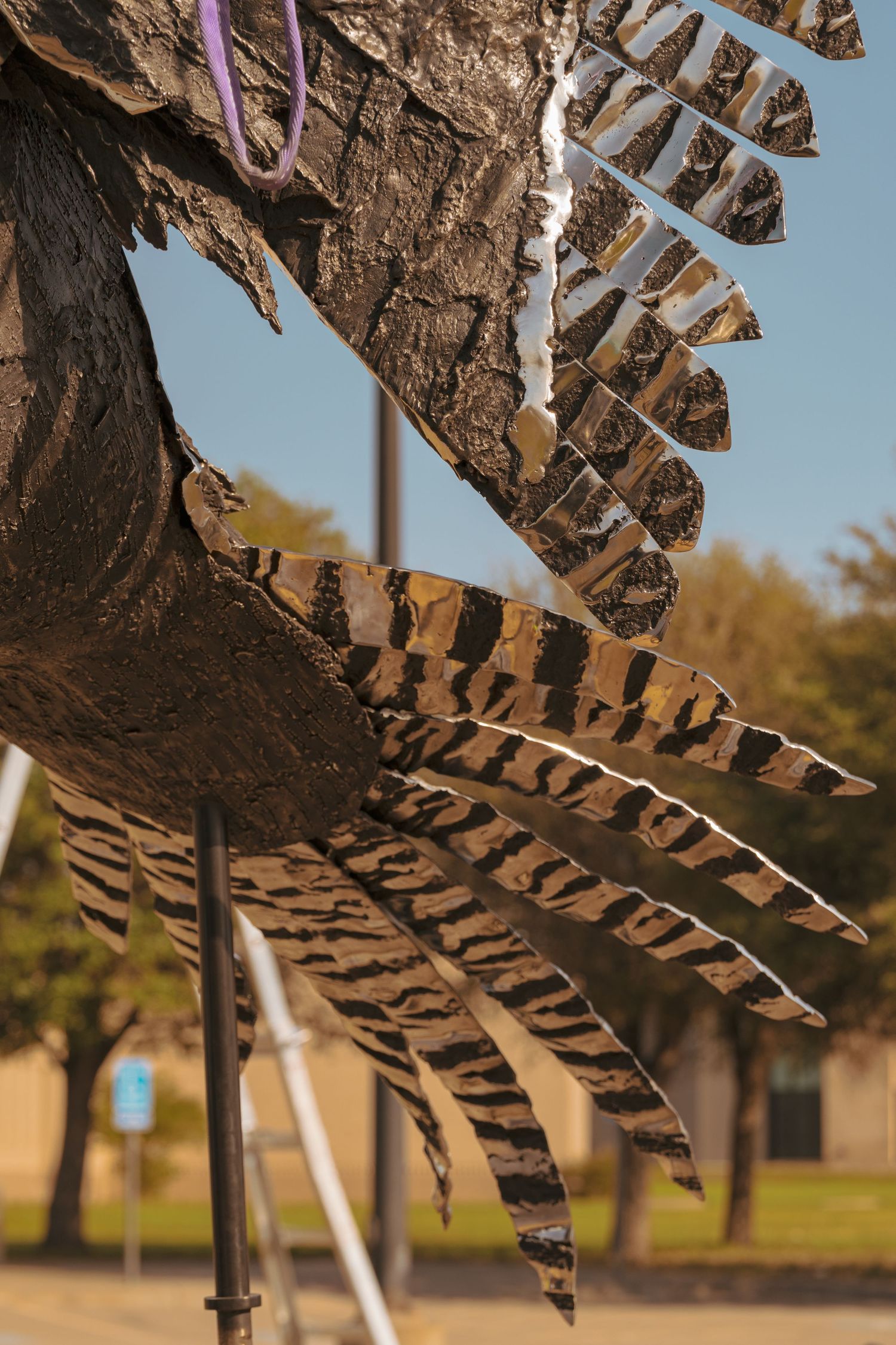 Detailed metal feathers of a large bird sculpture with striped pattern against a blue sky.