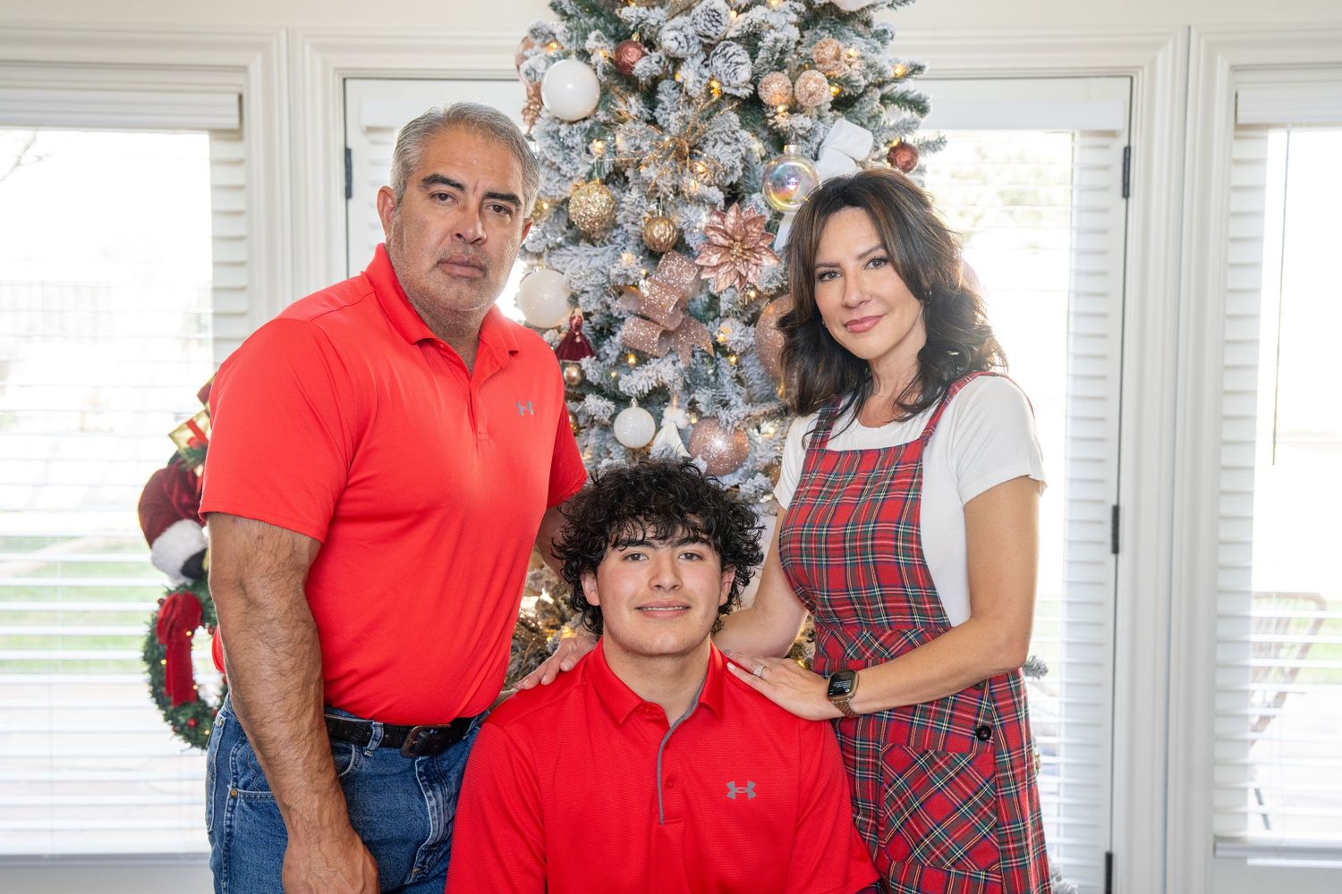 Family of three posing in front of a decorated Christmas tree, wearing festive red and plaid outfits indoors.