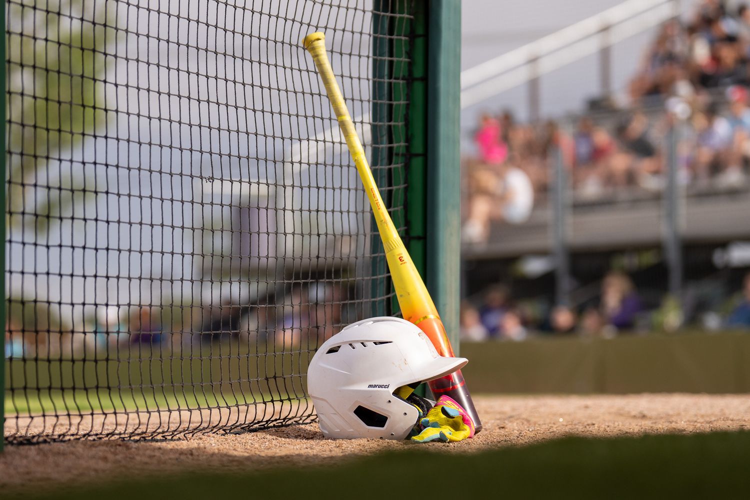 Yellow softball bat leaning against white batting helmet near backstop.