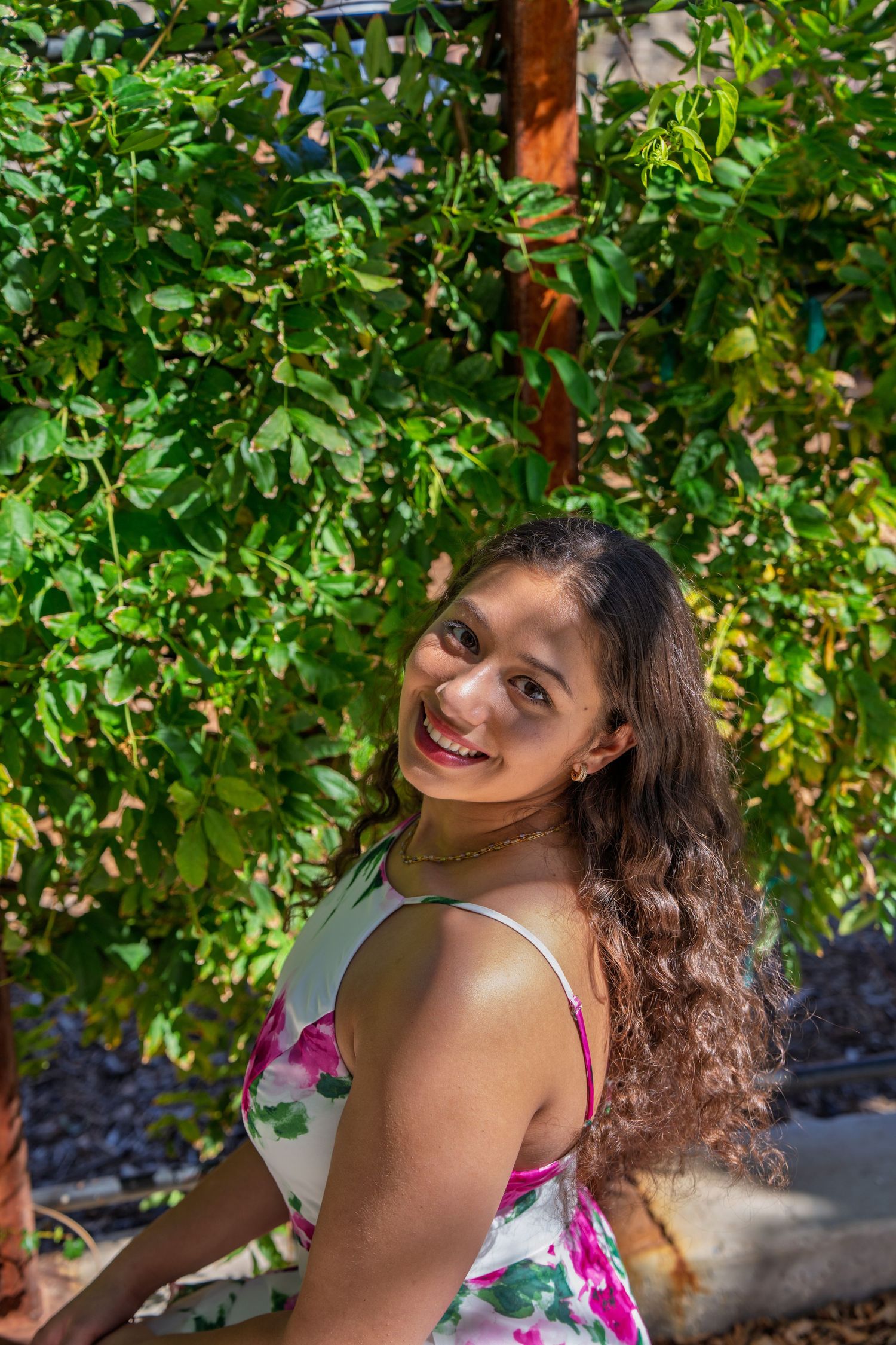 Smiling young Latina woman with curly hair in floral dress standing near green leafy plants outdoors.