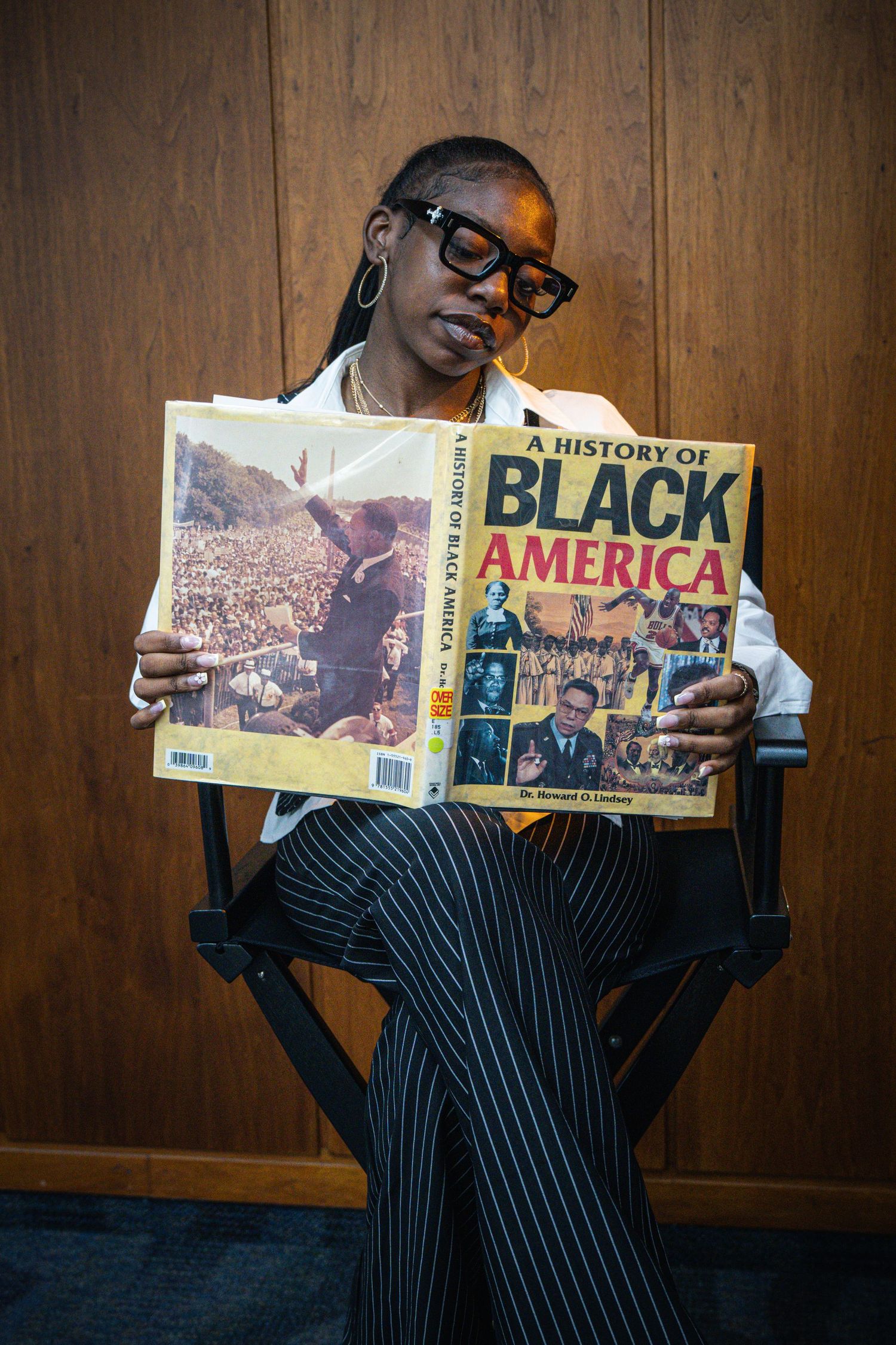 Black woman in pinstripe suit reads A History of Black America book while seated in a directors chair.