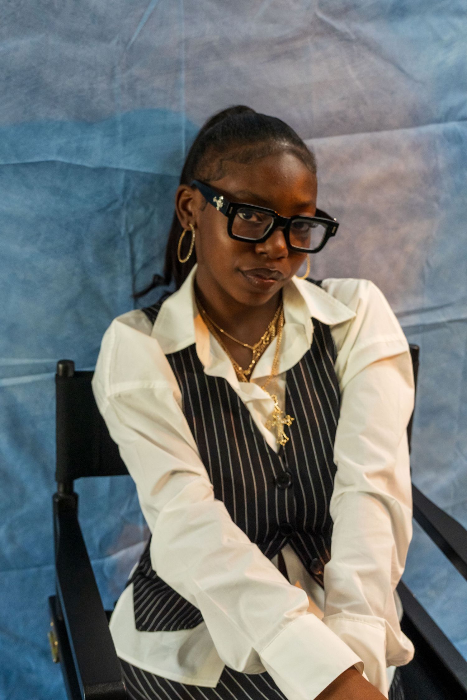 Young Black woman in pinstripe vest and glasses sits in director chair against blue backdrop.