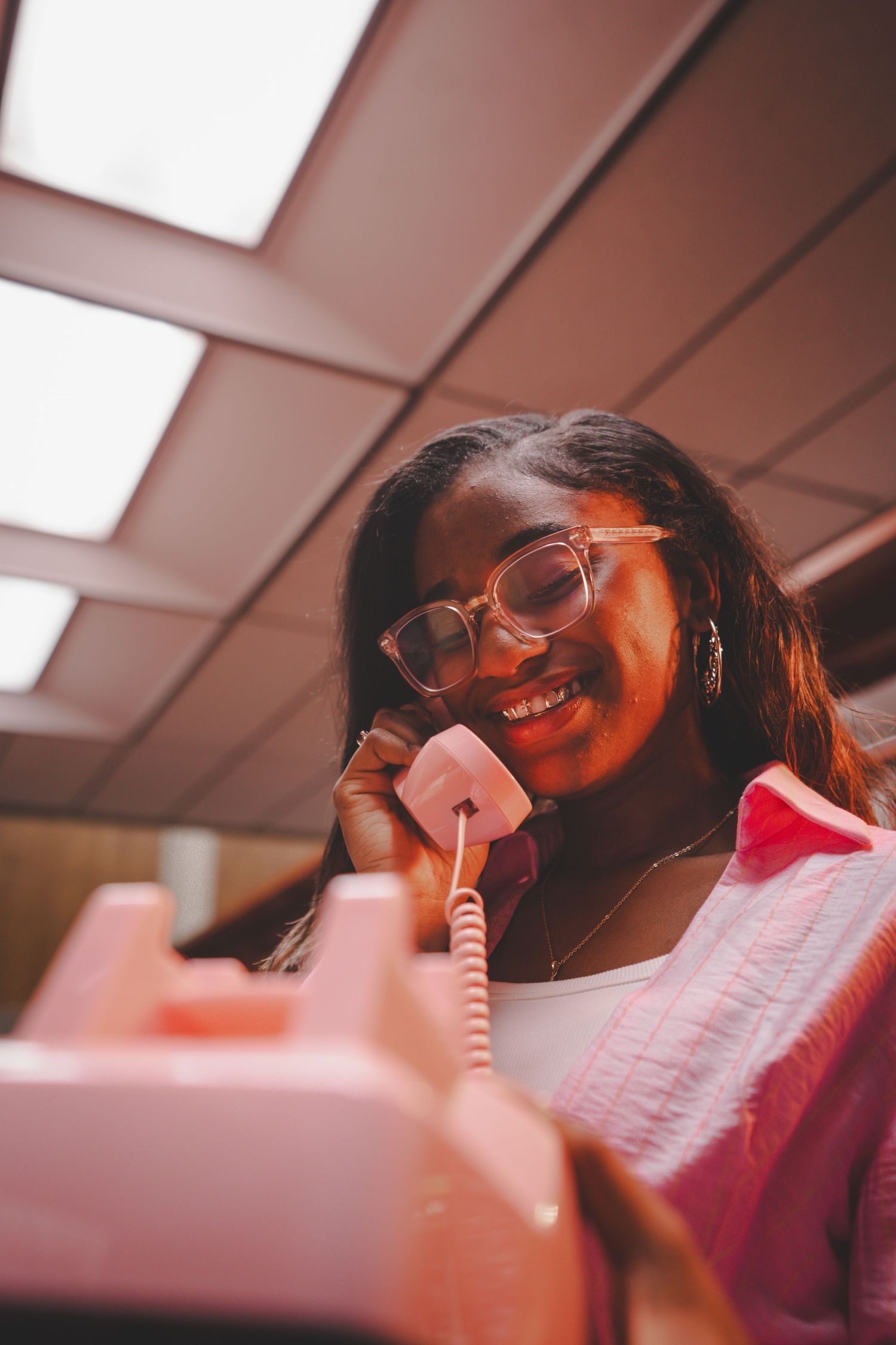 Young woman with glasses smiles while talking on retro pink telephone.