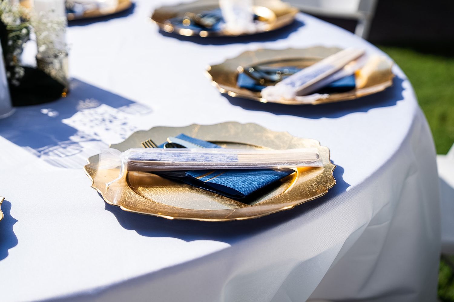 Elegant outdoor wedding table setting with gold plates, blue napkins, and silverware on a white tablecloth.