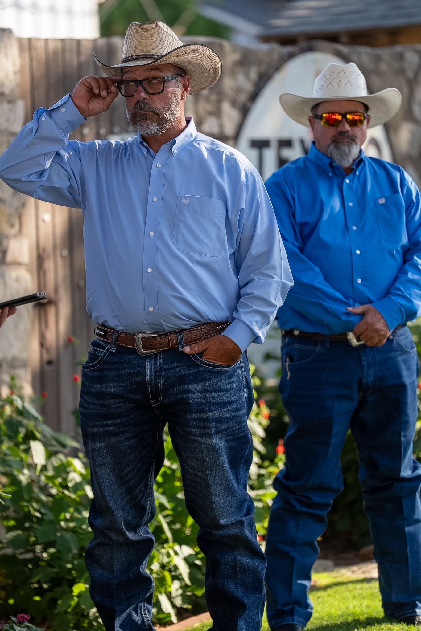 Two men wearing cowboy hats and blue shirts stand outdoors in a Western setting, one adjusting his hat.