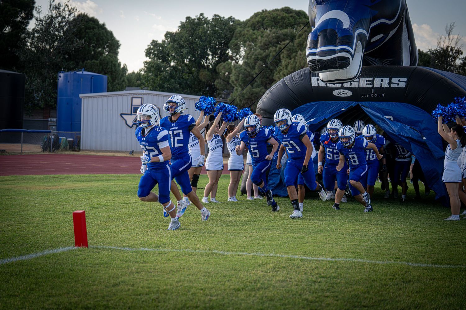 Youth football team in blue uniforms charging through inflatable tunnel onto field during game night.