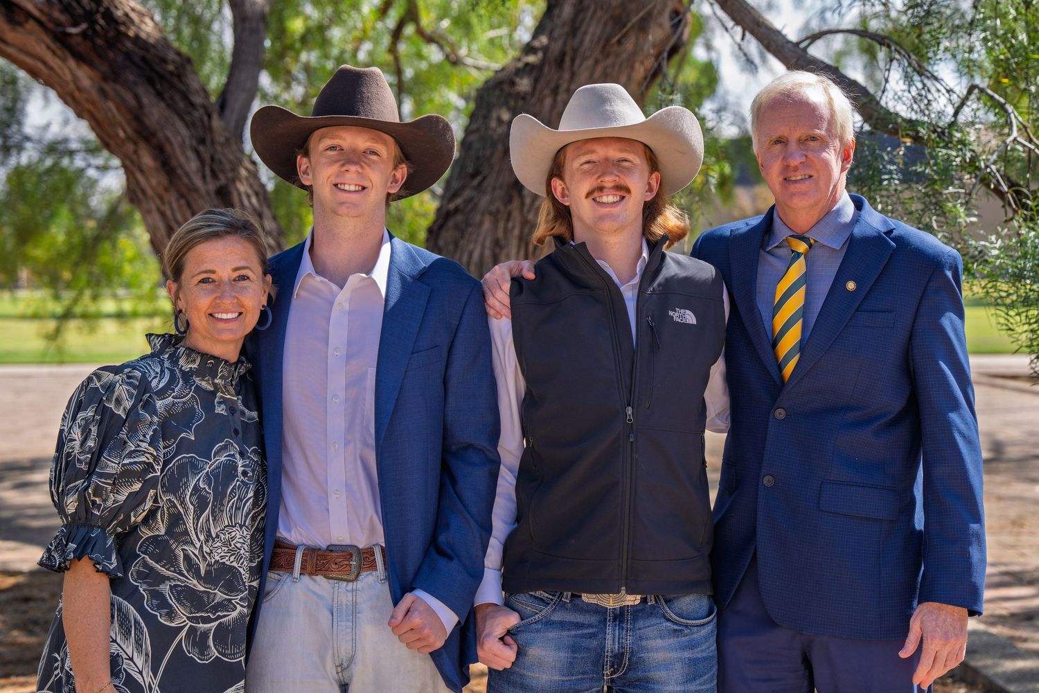 Four people smiling outdoors under trees, two wearing cowboy hats, dressed in smart casual and formal attire.