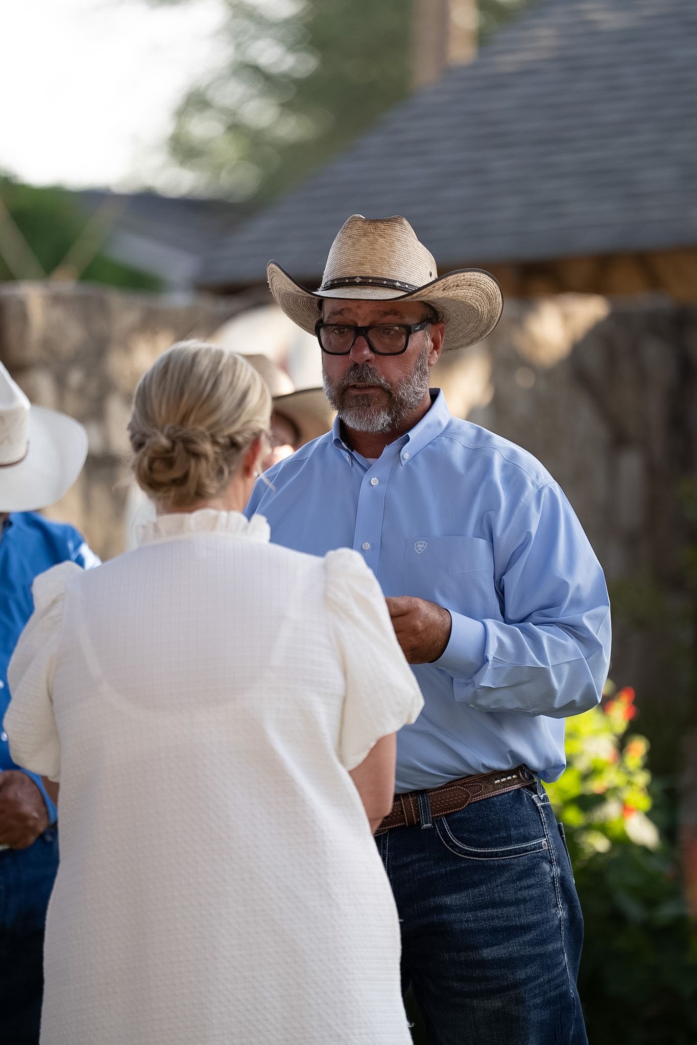 A bearded man in a straw cowboy hat and blue shirt converses with a woman in white at an outdoor gathering.