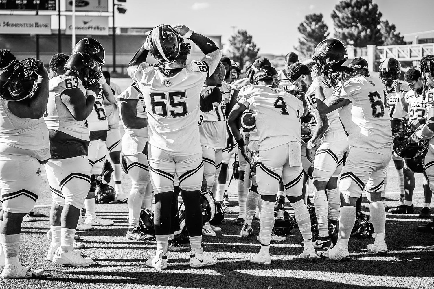 Black and white photo of football players in uniforms huddle on sideline during game.