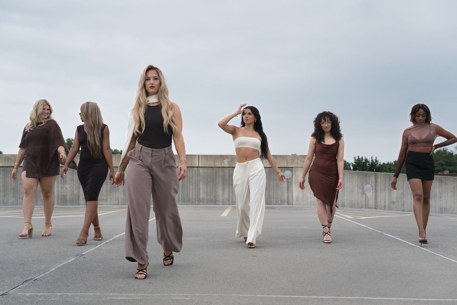 Seven diverse women in stylish outfits walking confidently in a parking lot under an overcast sky.