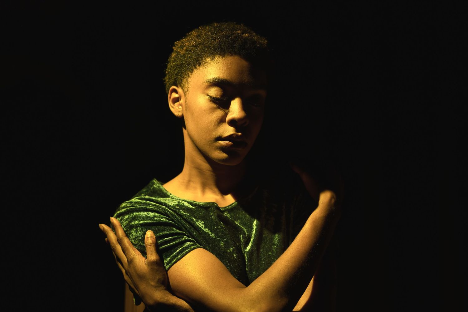 Young dancer in green costume performing with eyes closed on a dark stage.