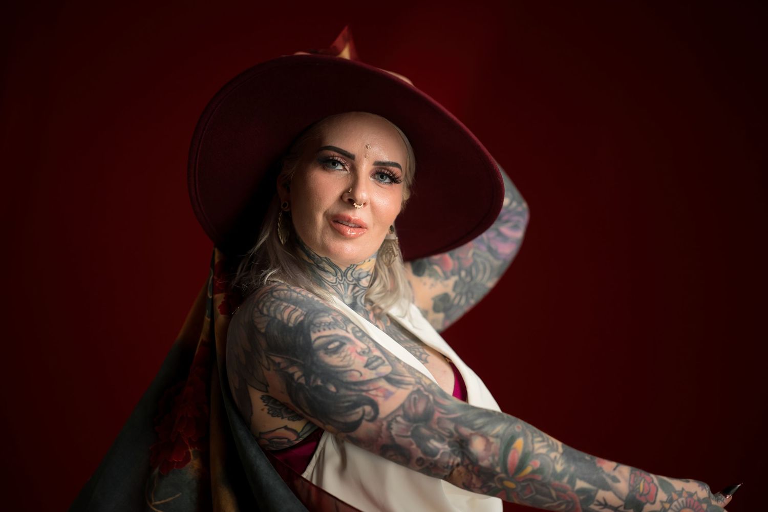 Tattooed woman in red wide-brim hat posing against dark red background.