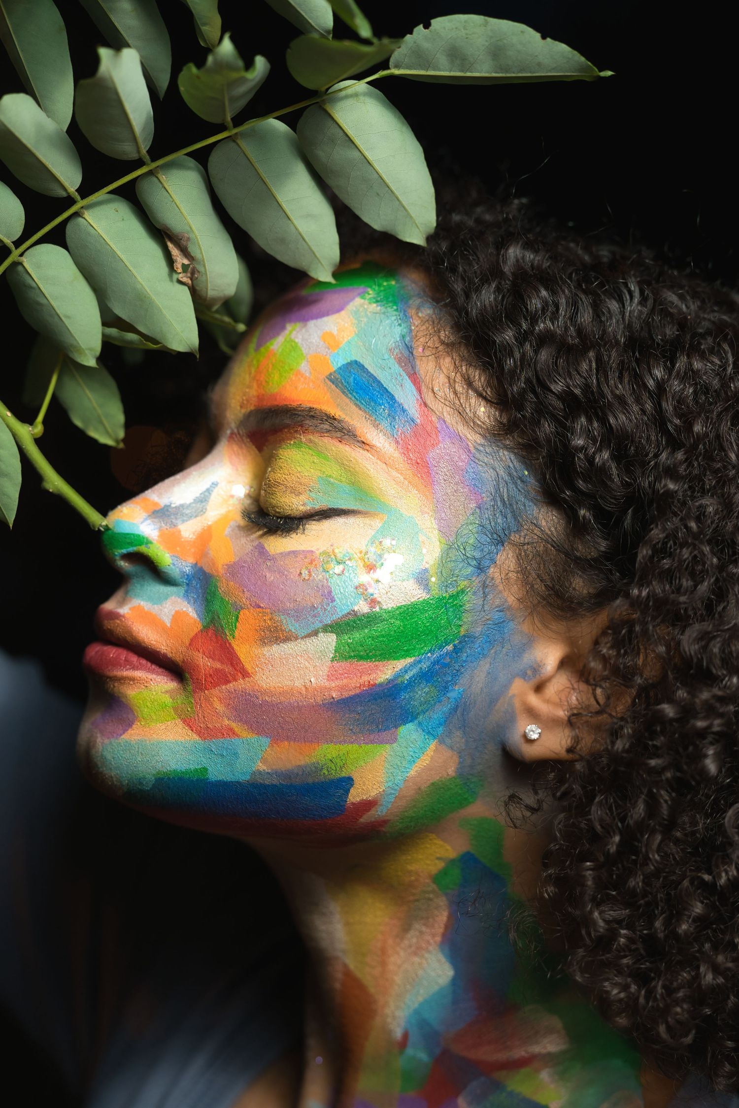 Woman with colorful face paint and curly hair posing with tropical leaves.