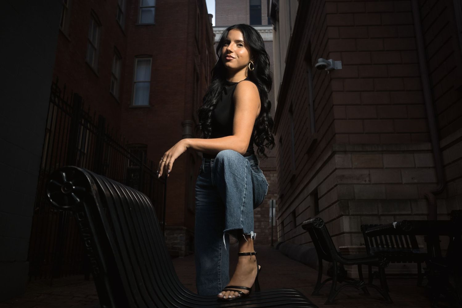 Woman in black top and ripped jeans posing on a bench in a dramatic urban alleyway.