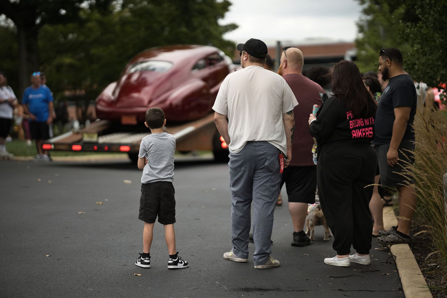 People watching a classic red car being transported on a flatbed truck in a parking lot.