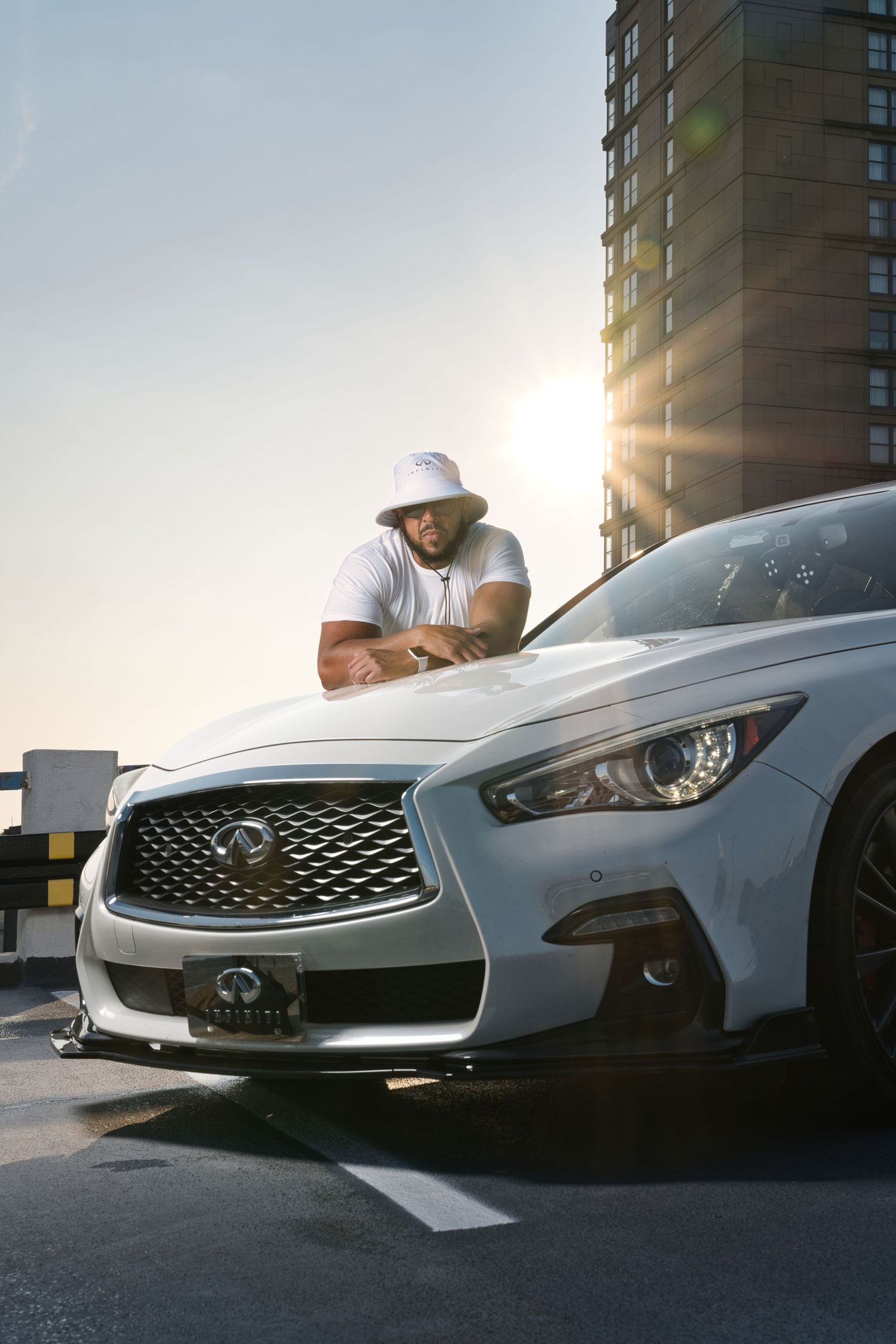 Man in white bucket hat leaning on silver Infiniti Q50 on rooftop parking garage at sunset.