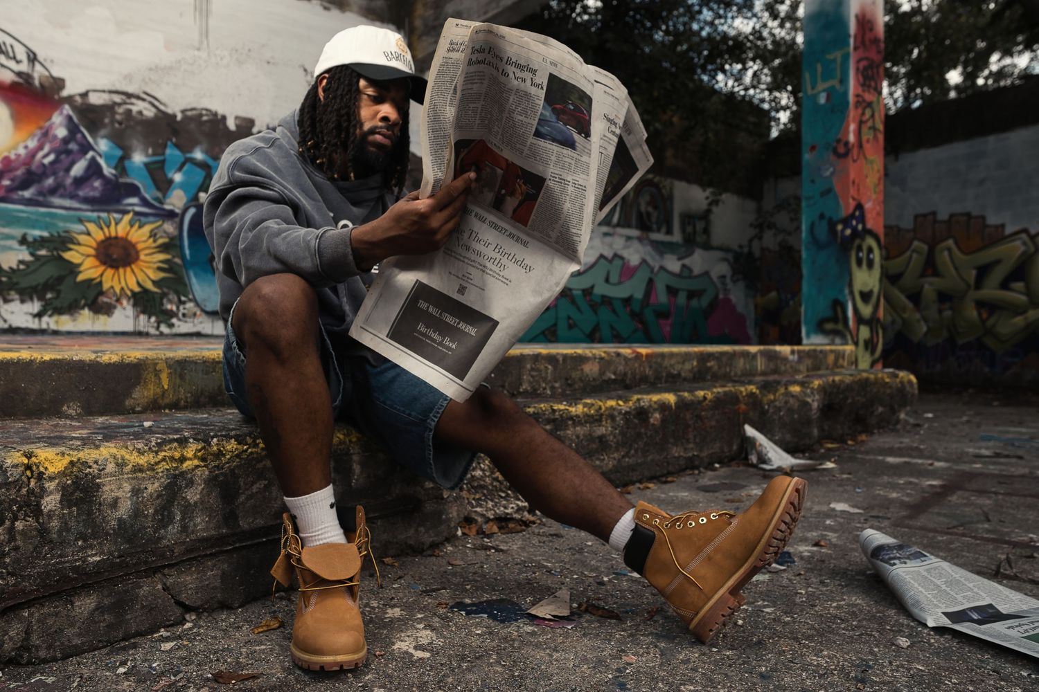 Man with dreadlocks reading a newspaper while sitting on steps near colorful graffiti wall outdoors.
