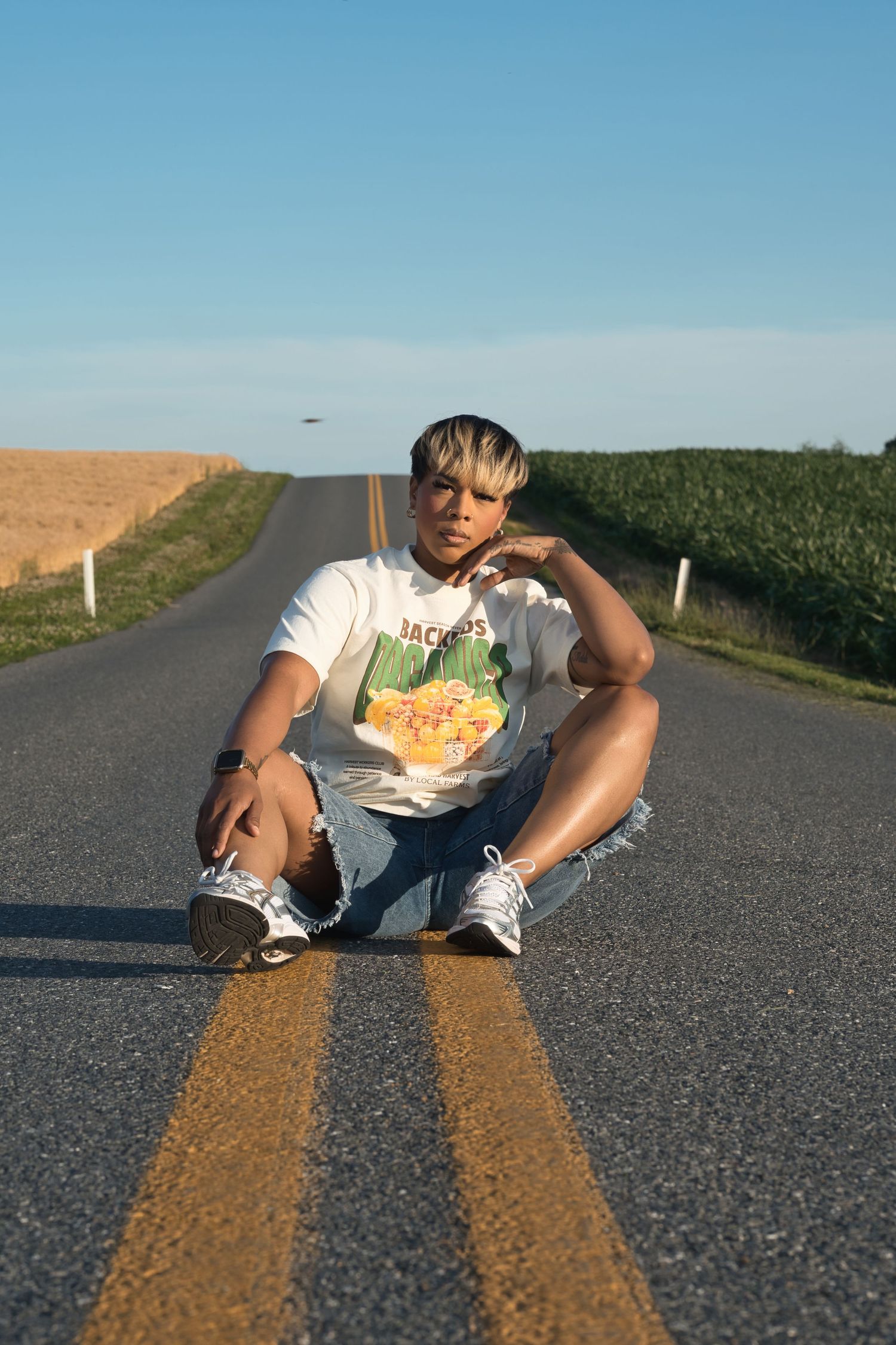 Young person with blonde highlights sitting on yellow center line of a rural road wearing a graphic tee and denim shorts.