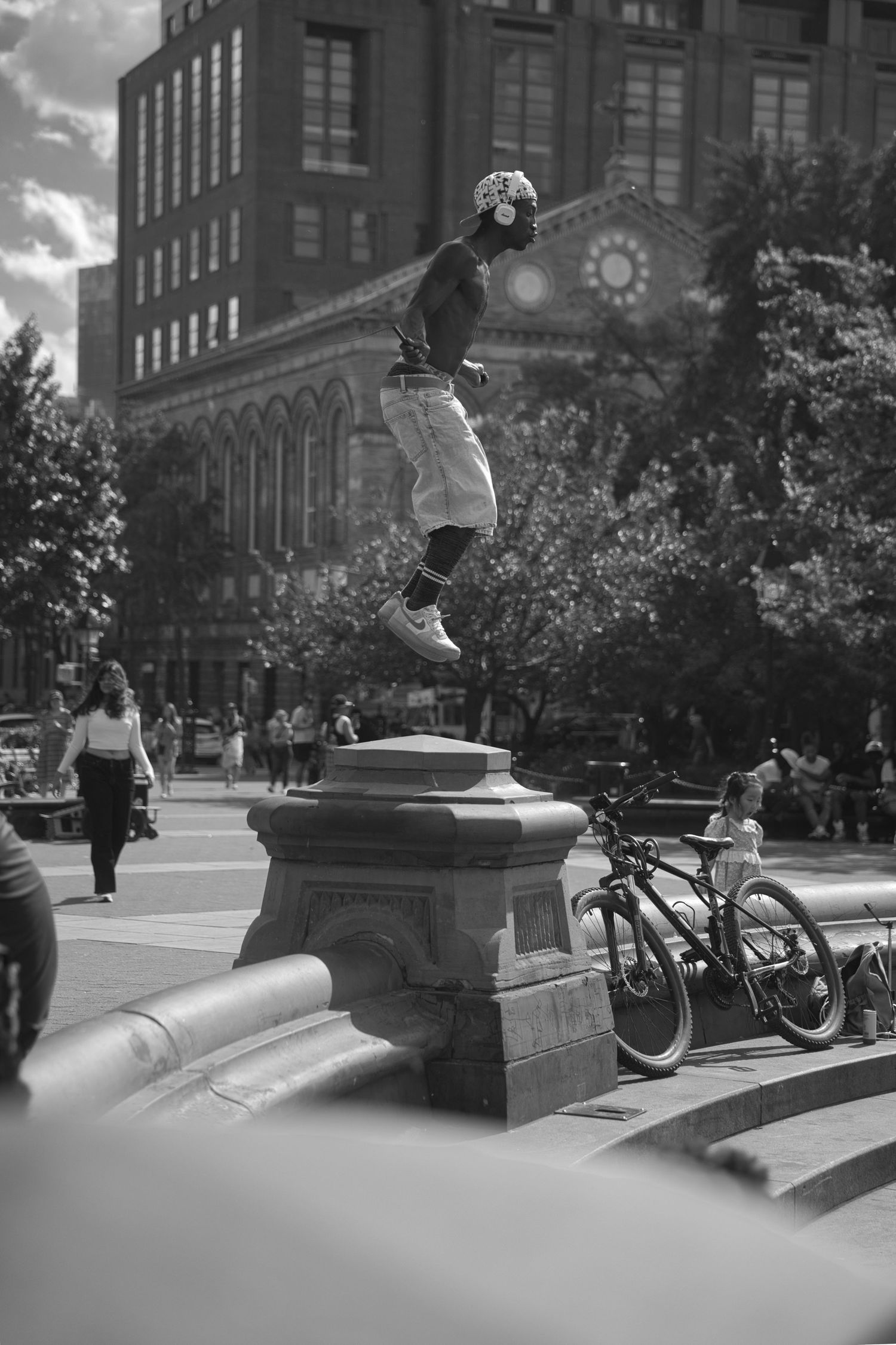 Black and white photo of a skateboarder jumping on a stone pedestal in an urban plaza with bikes and buildings.