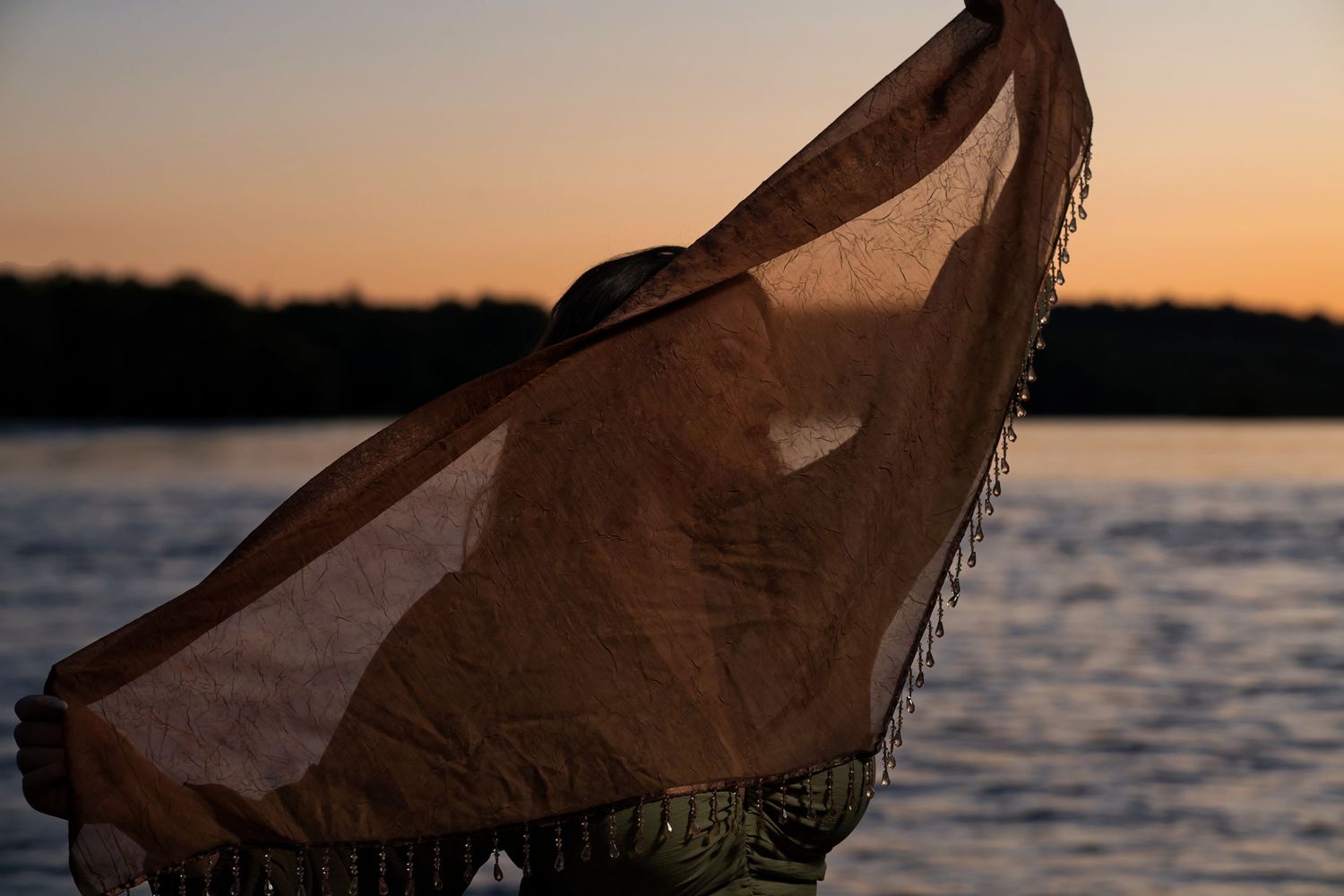 Person holding a sheer brown fringed scarf in the wind near a lake during a golden sunset.