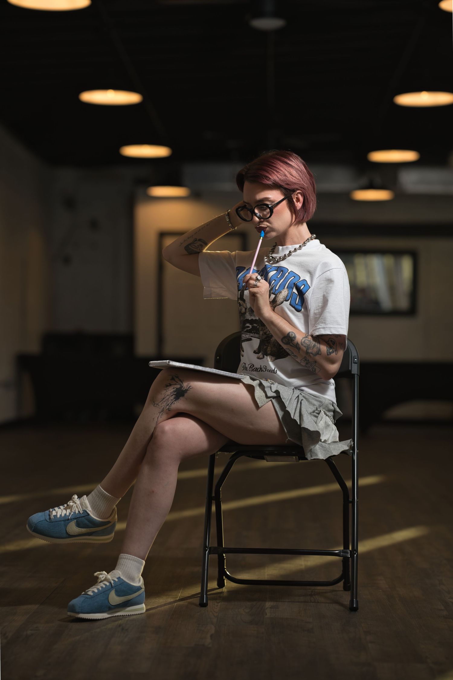 Tattooed woman with pink hair sits on folding chair holding pencil and sketchbook, wearing white tee, skirt, blue Nike sneakers.
