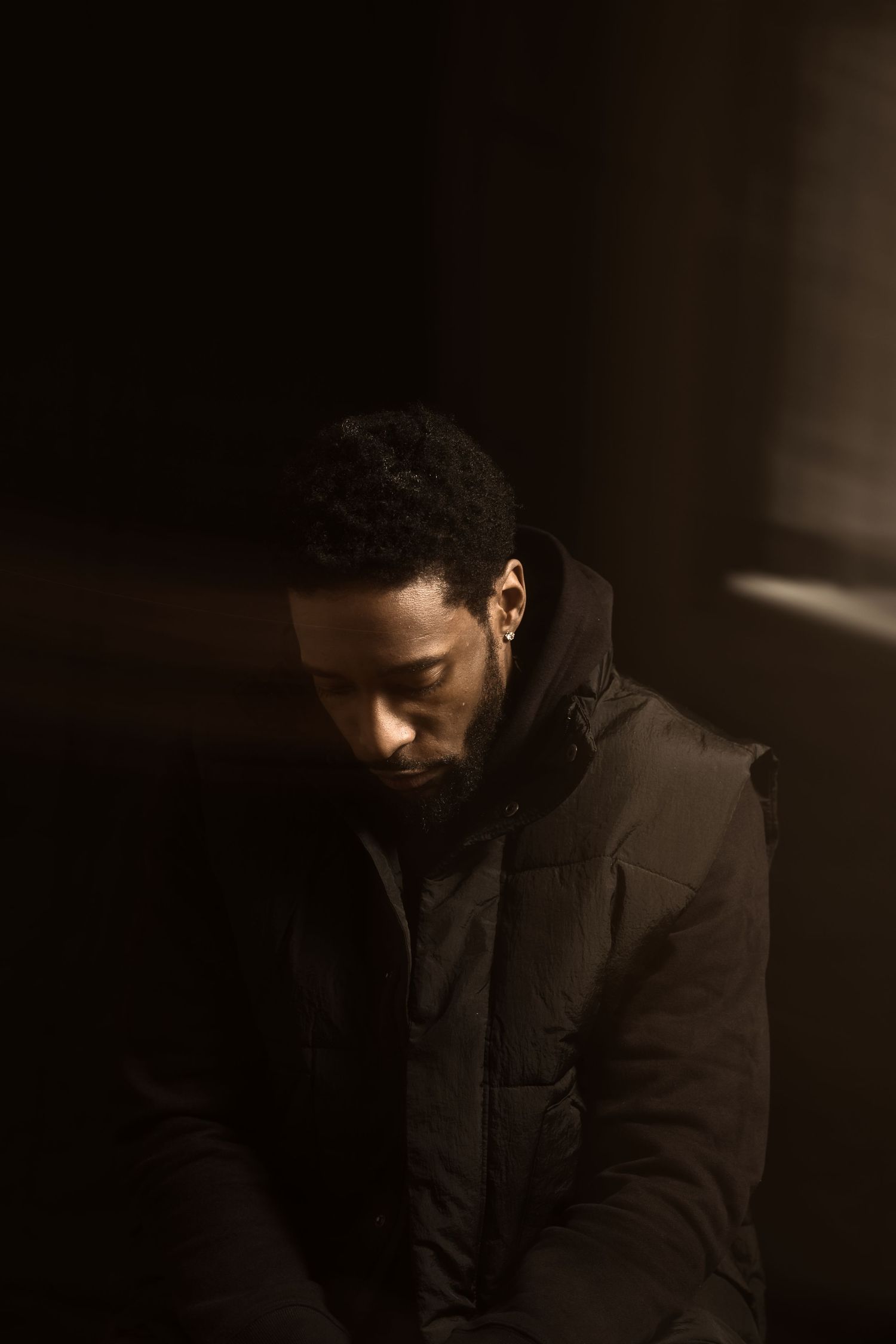 Young Black man in black puffer jacket looking down in dramatic moody lighting.