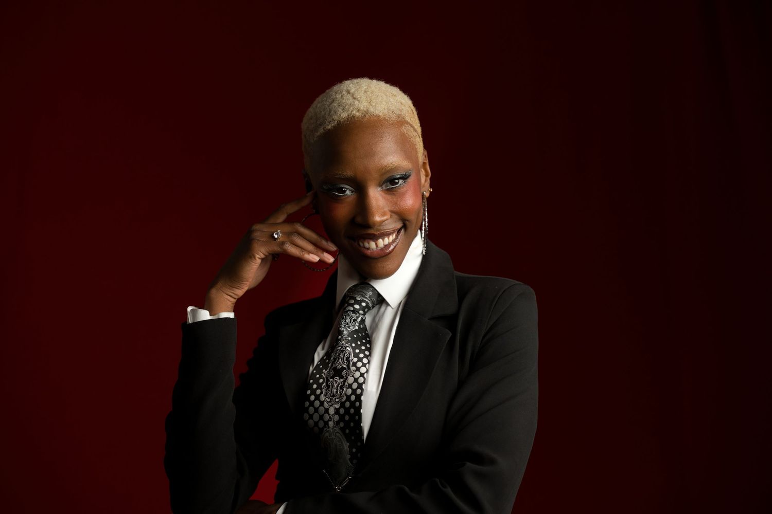 Smiling Black woman with blonde hair in black suit and patterned tie on red background.