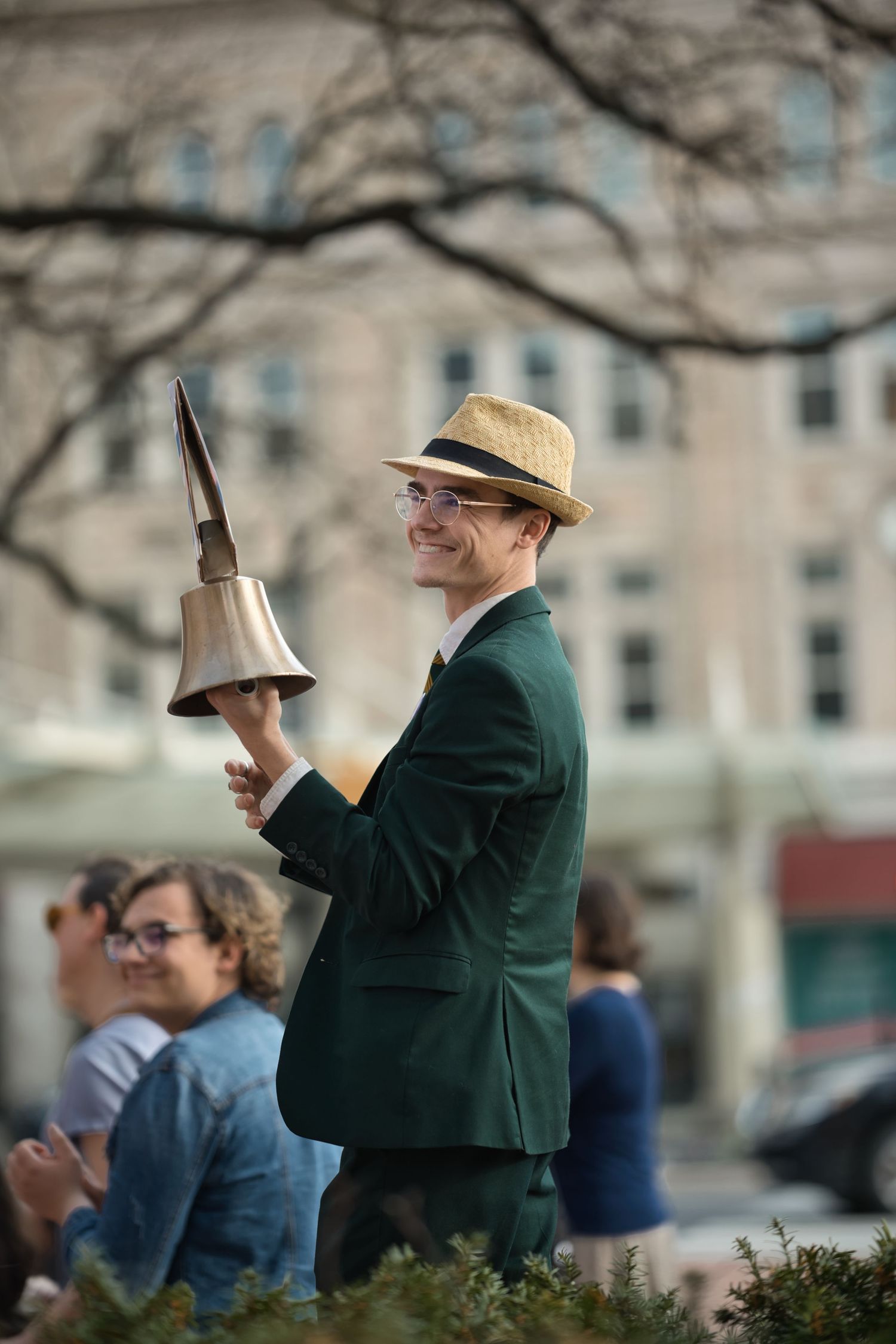 Smiling man in green suit and straw hat holding a brass bell outdoors at a public event.
