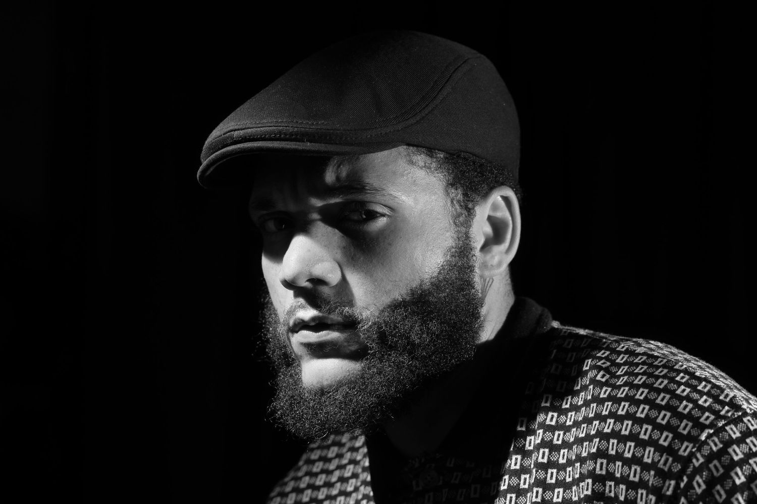 Black and white portrait of bearded man wearing flat cap against dark background.