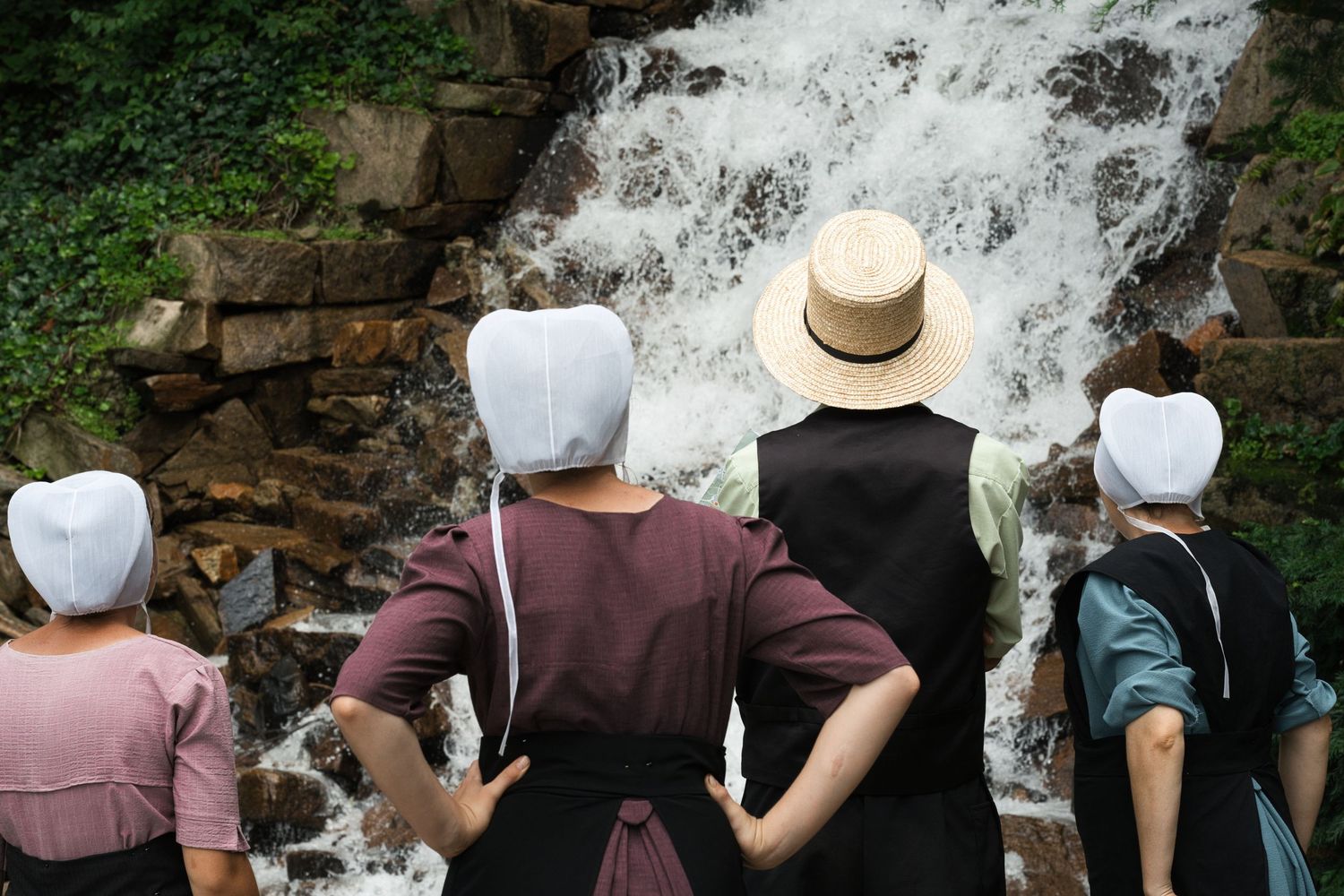 Amish women in traditional dress and bonnets viewing a rushing waterfall outdoors.