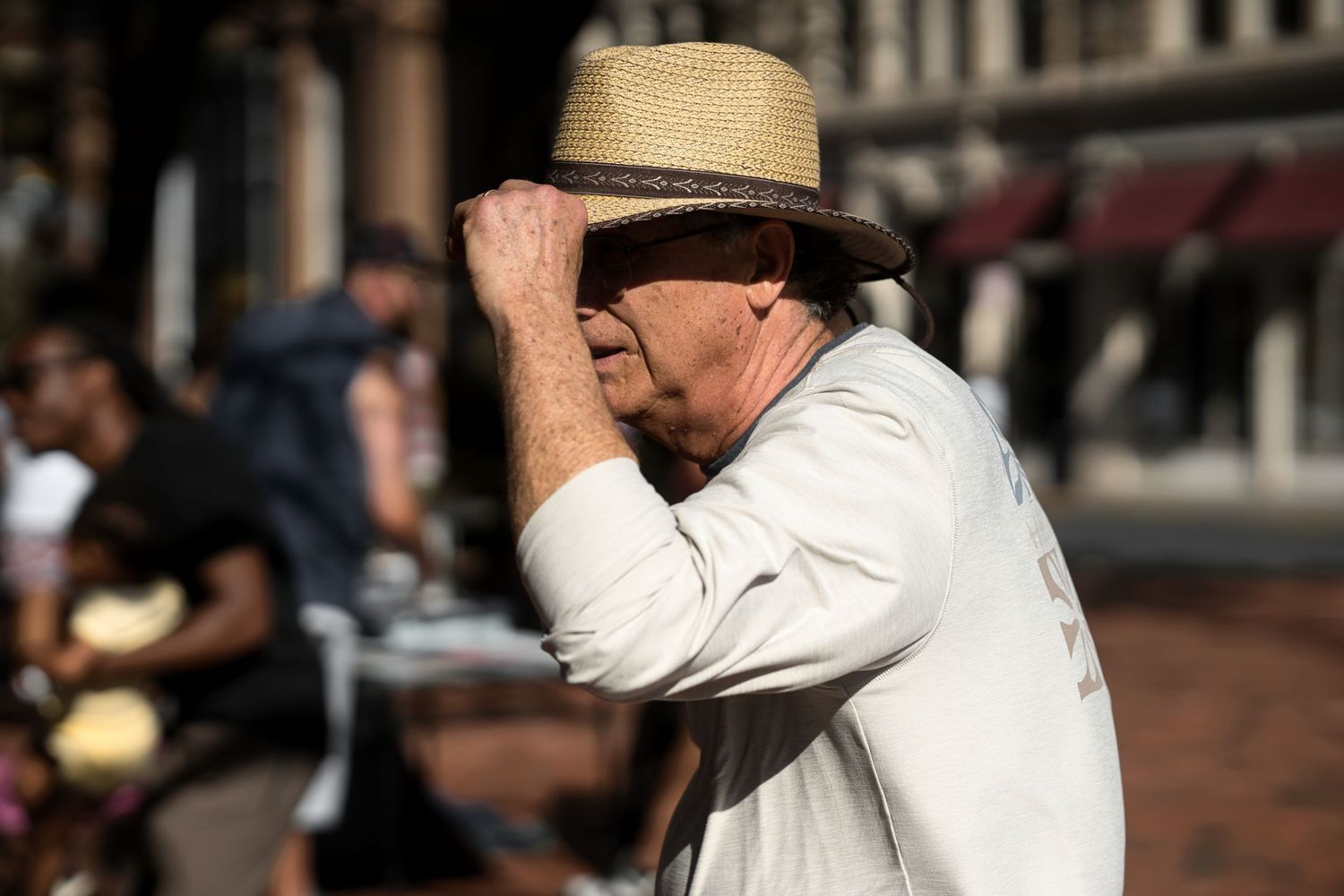 Elderly man shielding his eyes from the sun while standing on a busy street.