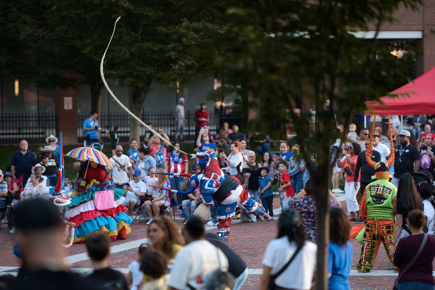 Colorful cultural festival performers dancing outdoors at dusk with crowd of spectators gathered in an urban plaza.