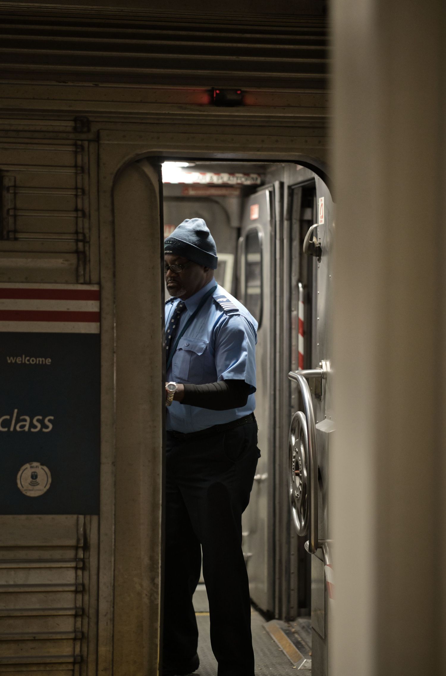 A train conductor in a blue uniform and cap stands in the doorway between two train cars at a station.