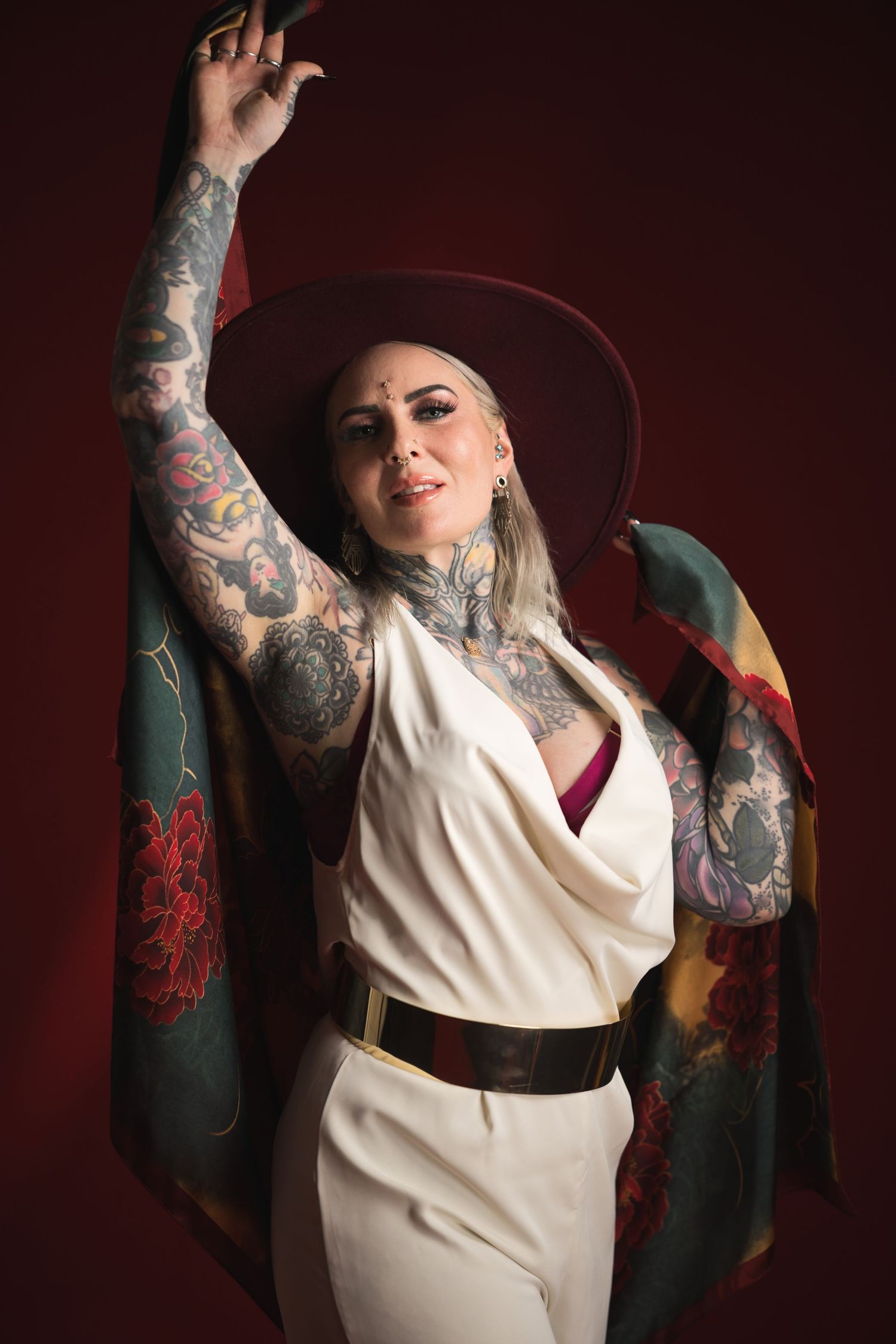 Tattooed woman in white jumpsuit, burgundy wide-brim hat, floral shawl, posing confidently against dark red background.