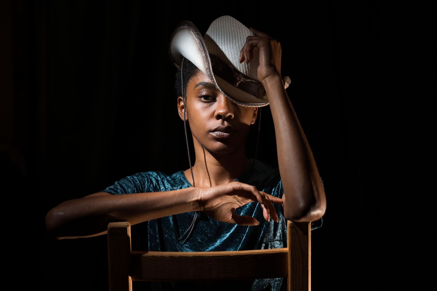 A Black woman poses elegantly in a chair, holding a white hat near her face against a dark background.