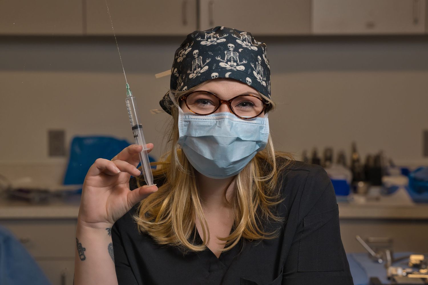 Female healthcare worker in scrub cap, mask, and glasses holding a syringe in a medical setting.