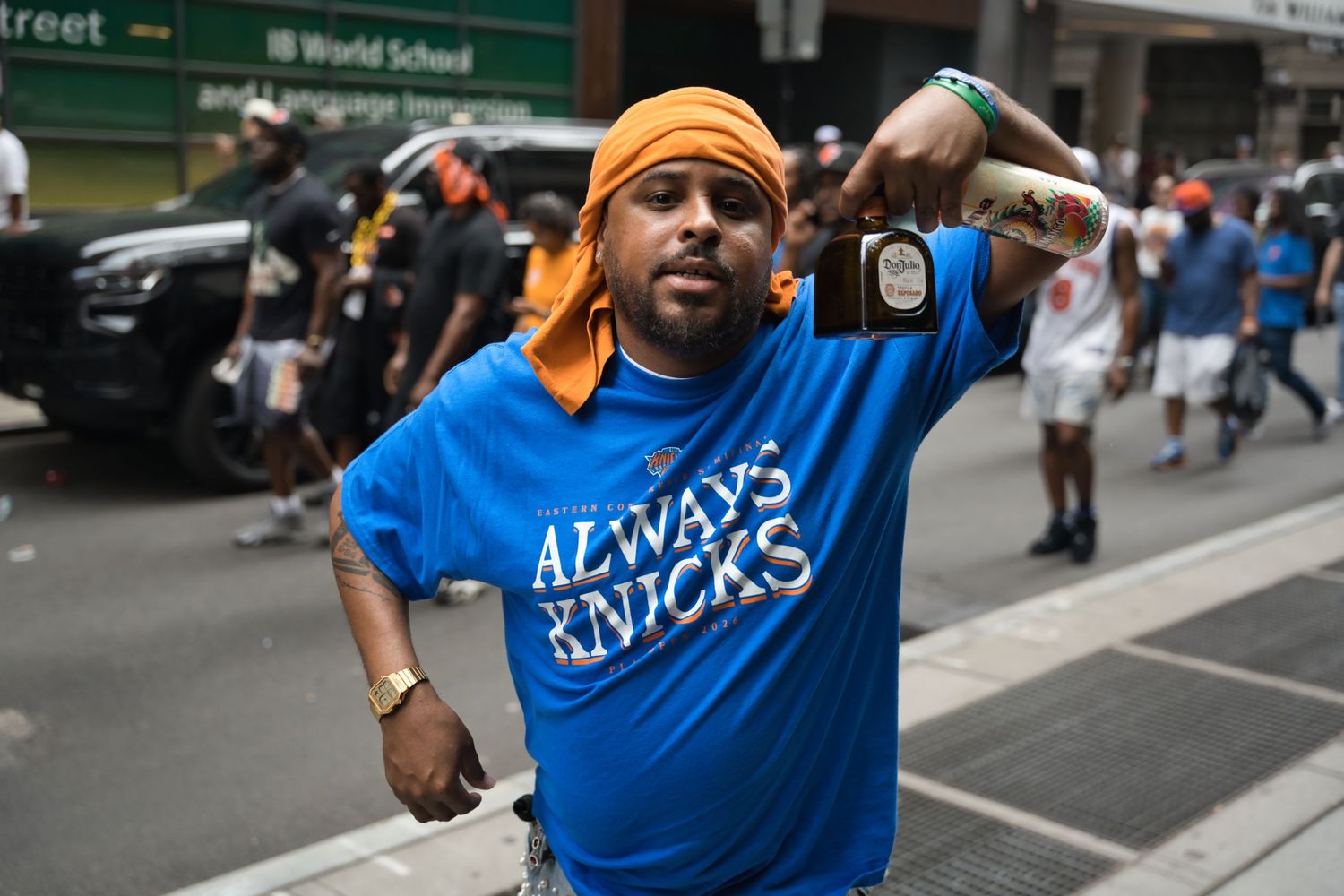 Man wearing an Always Knicks blue t-shirt and orange bandana holding a speaker at an outdoor street event.