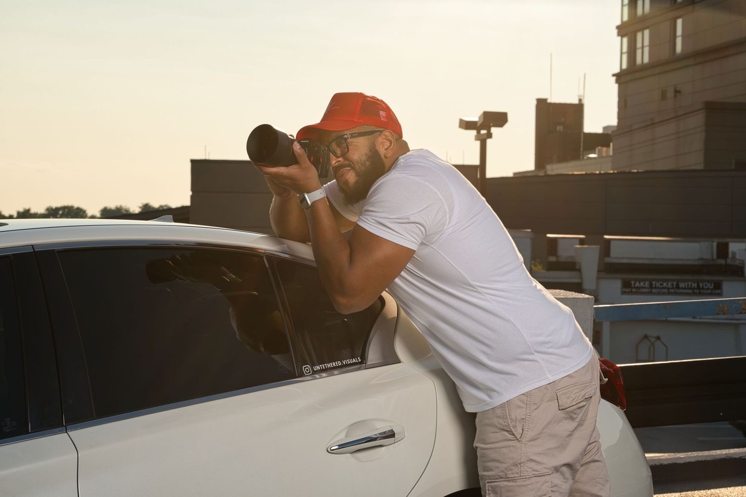 Bearded man in white tee photographing white car with telephoto lens at golden hour.