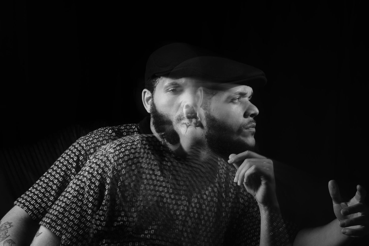 Black and white portrait of a bearded man in a cap exhaling smoke, looking thoughtfully to the side.