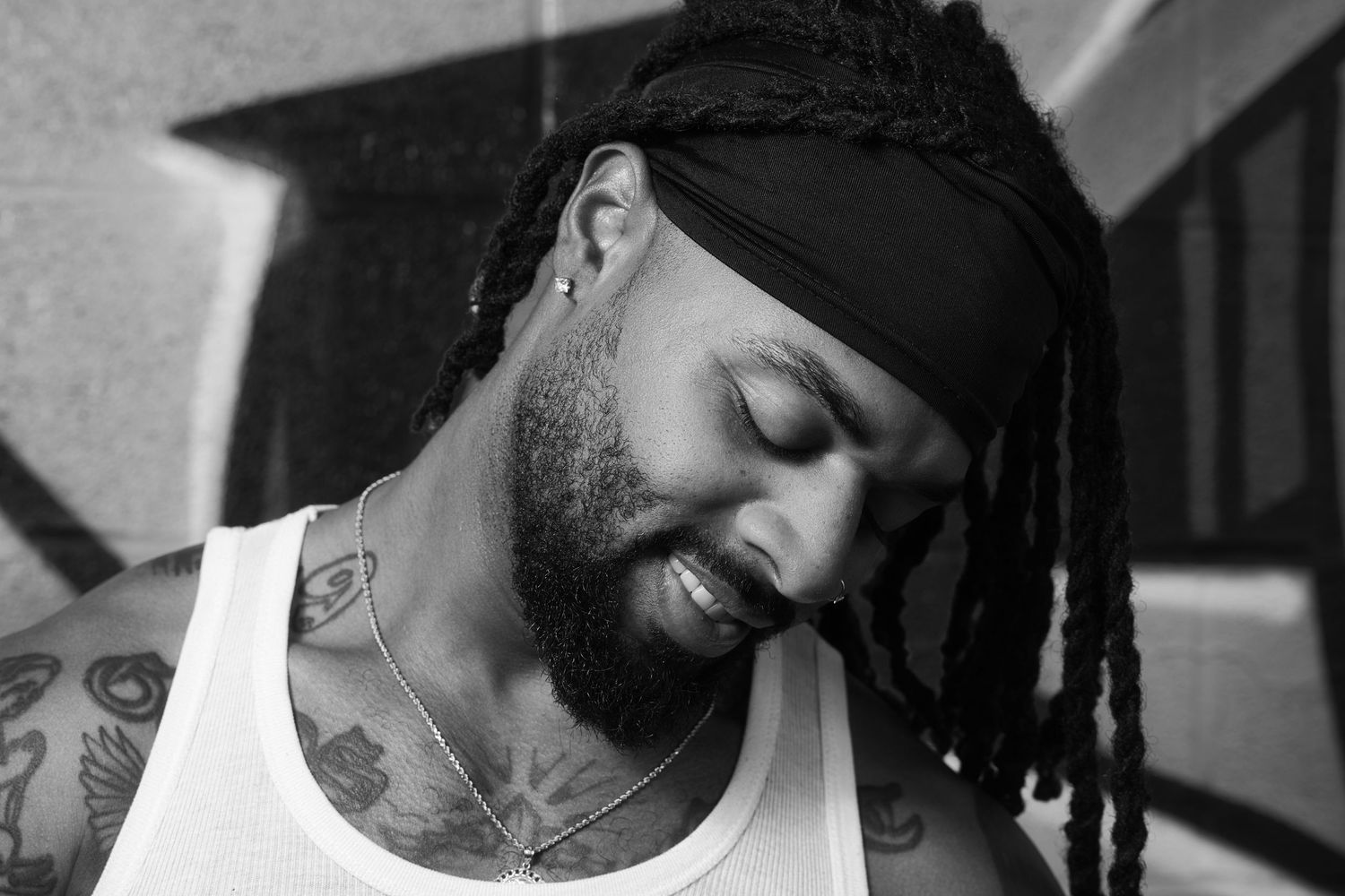 Black and white portrait of tattooed man with dreadlocks and headband looking down.
