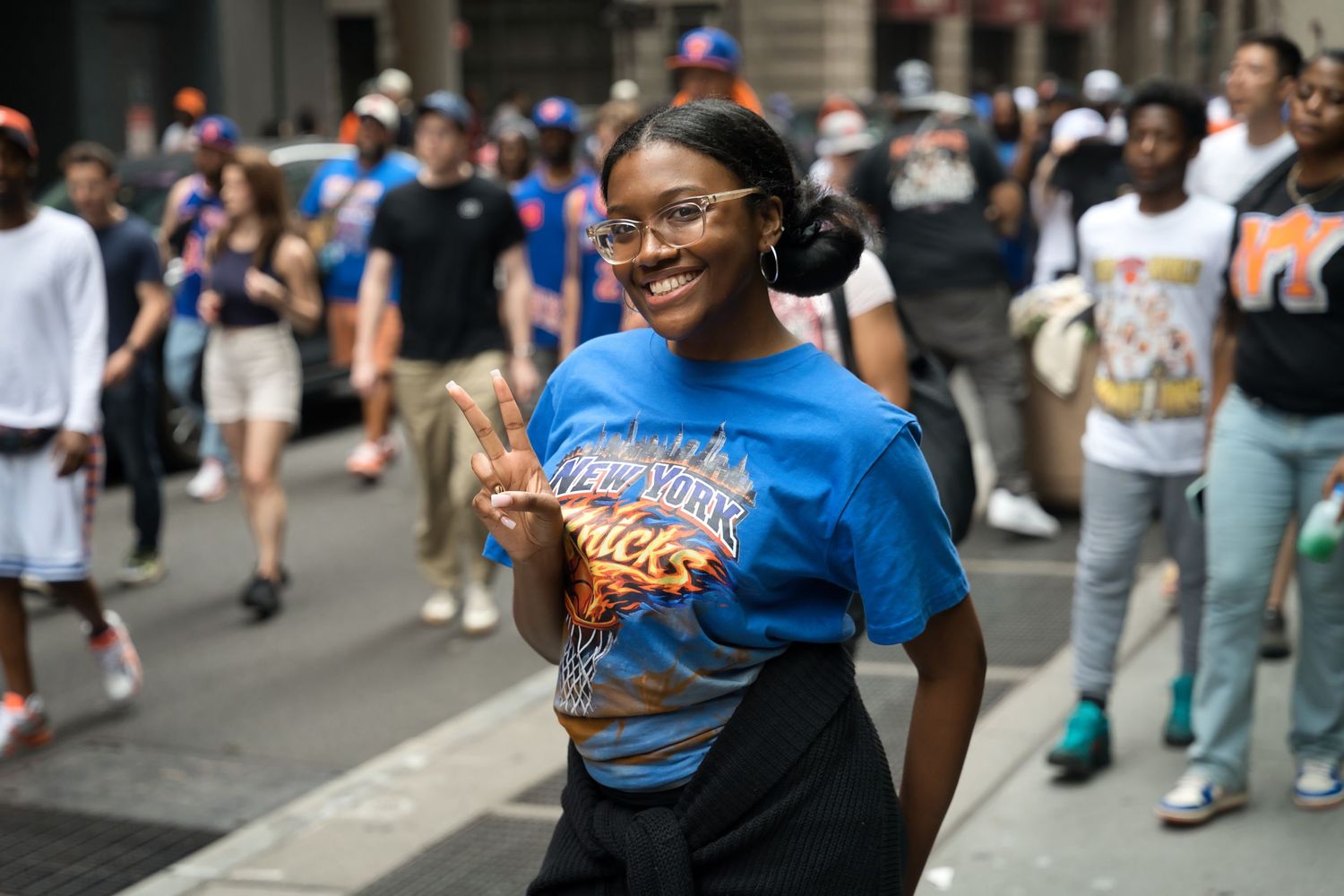 Young woman in blue New York Knicks t-shirt flashing peace sign at outdoor street parade.