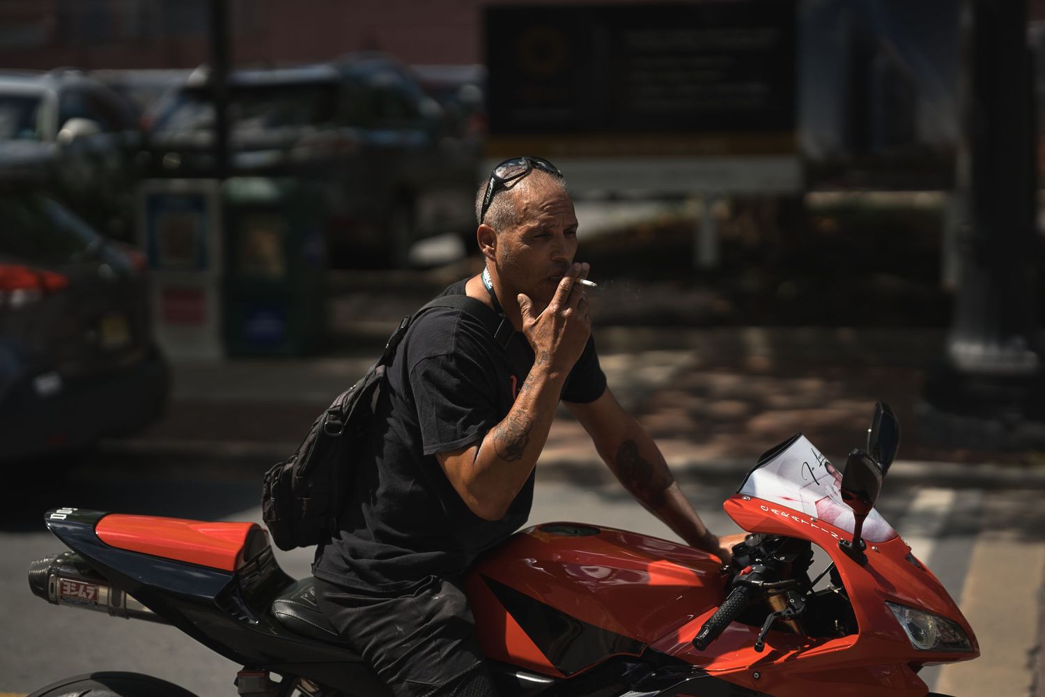 Man in black shirt smoking a cigarette while leaning on a red motorcycle outdoors on a sunny day.