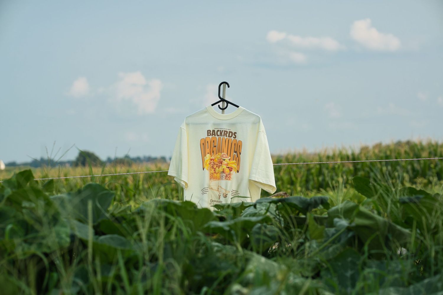 Cream colored graphic t-shirt hangs on hanger in lush green farm field under blue sky.