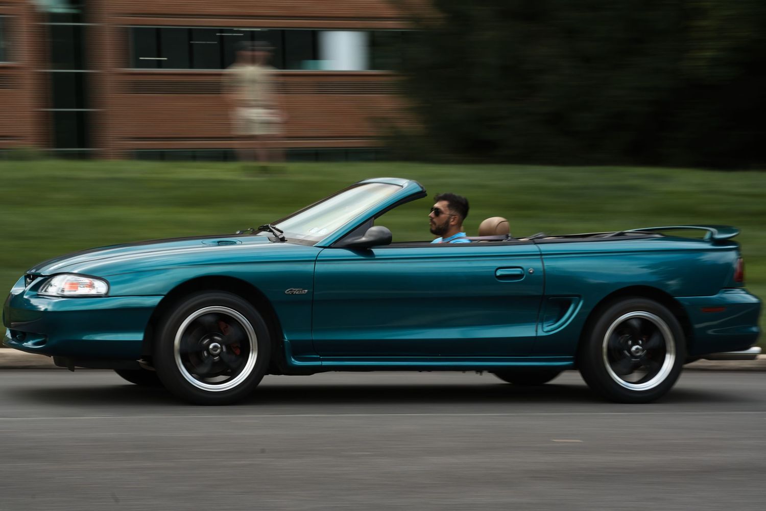 Teal Ford Mustang convertible driving on a road with two passengers, featuring white-wall tires and a spoiler.
