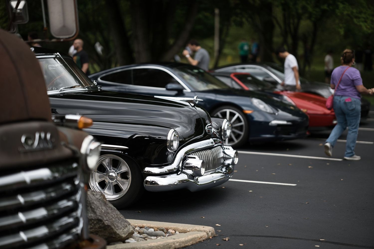 Vintage black custom car with chrome bumper parked alongside a Porsche and red sports car at an outdoor car show.