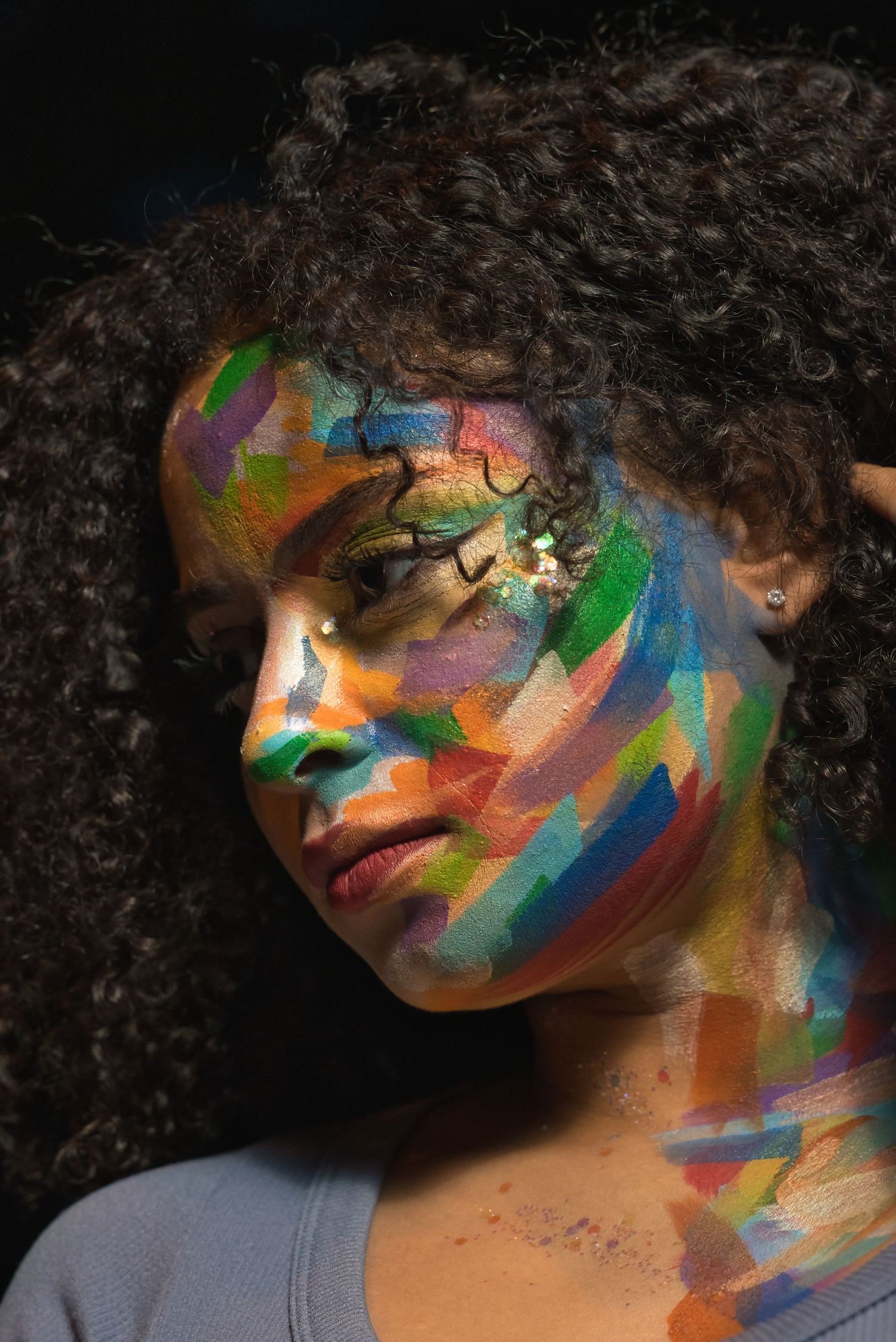 Close-up portrait of a woman with vibrant multicolored face paint and curly hair in dark lighting.