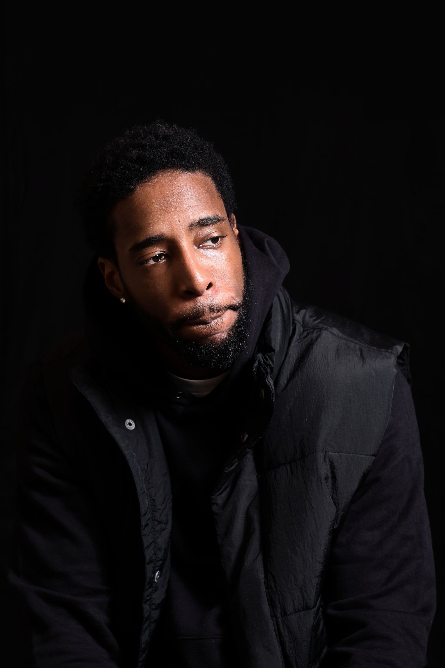 Young Black man in black puffer jacket gazing at camera against black background.