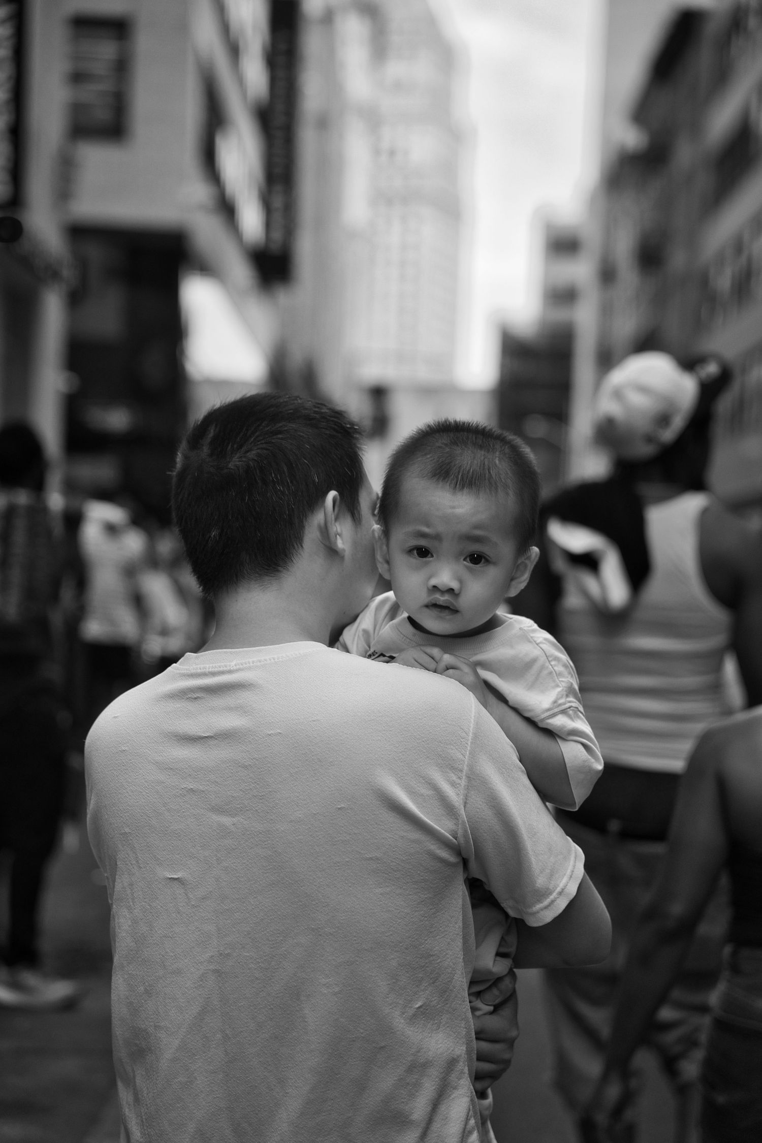 Black and white street photo of a man carrying a young toddler in a busy city.